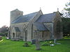 Gray stone building with square tower at left hand end. In the foreground are gravestones on grass.