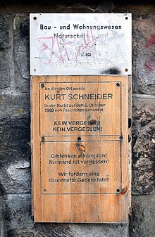 Eine Holzplatte in die schwarze Buchstaben gebrannt wurden, befestigt auf einer Steinwand. Darauf steht: "An diesem Ort wurde Kurt Schneider in der Nacht auf dem 06.10.1999 von Faschisten ermordet. Kein Vergeben! Kein Vergessen! Gedenken erkämpfen! Niemand ist vergessen. Wir fordern eine dauerhafte Gedenktafel!"