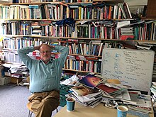 He is leaning back from a table with two cups of coffee/tea on it, with an enormous wall of books piled haphazardly on shelves behind him. There is also a whiteboard shown with equations.