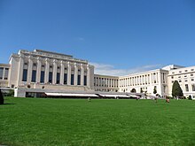 Un camino conduce más allá de un césped bien cuidado hasta un gran edificio rectangular blanco con columnas en la fachada.  Dos alas del edificio están retranqueadas desde la sección central.