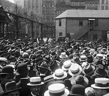 Ein Platz voller Menschen mit Hüten, in der Mitte des Bilds Emma Goldman, die mit ausgestrecktem Zeigefinger redet.