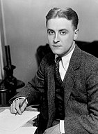 Photographie de Fitzgerald vers 1921. Il regarde la caméra alors qu'il est assis à un bureau avec un crayon dans sa main droite.  Il porte un costume sombre et une cravate noire à pois.