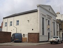 Three-quarter view of a wide, long stuccoed building, with the side wall more prominent. This is mostly a blank wall, but there are three windows towards the rear. The façade has three tall windows tapering from bottom to top; the central window has a small pediment and is shorter than the others to accommodate an entrance door. Above the roofline is a large pediment with louvred circular opening.