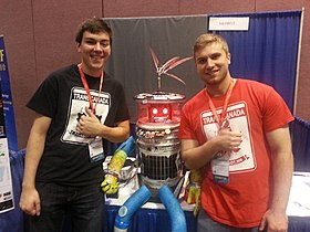 Dos personas de pie sonriendo enmarcan un robot humanoide sentado en una mesa.