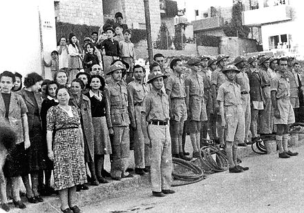 Jewish Civil Defense group in Jerusalem in 1942. The group served as ARP Fire Wardens, equipped with water hoses and buckets, some wearing FW (Fire Watcher) Brodie helmets. Men are in uniform while women wear plain clothes. Composer Josef Tal stands next to the woman with a black sweater.