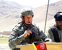 un uomo che indossa un'uniforme da combattimento dell'esercito che parla su un podio in primo piano, un uomo afgano in piedi a destra sullo sfondo.