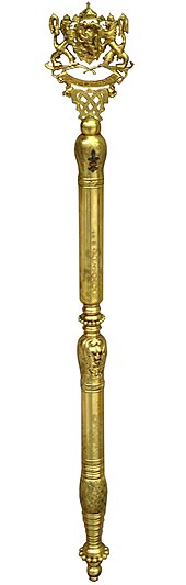 Photo verticale d'un sceptre doré décoré par deux lions, sur fond blanc.