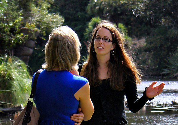 Two women talking to each other. Notice the woman in blue has an arm next to her body, whilst the other uses hers to gesticulate; both are signs of body language