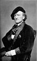 A black-and-white photograph of a man looking at the viewer while wearing a dark beret and a dark jacket and sitting on a chair, all in front of a light background