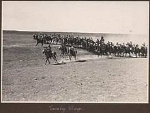 A large group of men on horses charging across a dusty plain, waving weapons.