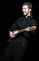 Chris Martin vistiendo un traje negro y tocando una guitarra eléctrica marrón.