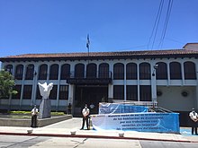 This is a picture showing the building of the Constitutional Court of Guatemala. It shows a two story building colonial Spanish type, with multitude of windows and a reddish tile roof, with a background of a blue sky on top. In the foreground there is a white sculpture depicting Pegasus, next to a concrete pathway leading to the entrance of the building. There are three police standing on the sidewalk apart from each other, in guard duty. There is also a couple of large banners between two of the police in the right of the picture, about the mission of the Court and recommending people to respect the integrity of the building