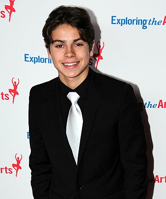 A mid shot of a teenage boy with dark brown hair parted to the sides, wearing a black shirt and blazer with a white tie. He is posing at a press event and smiling, looking directly towards the camera. Behind him are hoarding signs with names of commercial sponsors.