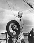 A man wearing a garrison cap and windbreaker paces on the deck of a ship.