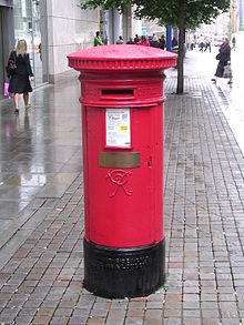 Rød sylindrisk boks med svart underlag.  En liten gylden plakett henger i midten av esken.