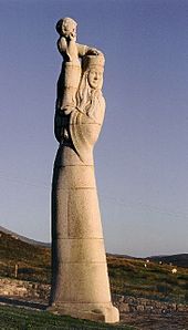 Een groot stenen beeld van een lange en slanke vrouw bij een veld.  De vrouw heeft lang haar en draagt ​​een hoed en draagt ​​een kind op haar schouders.