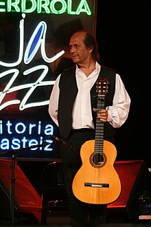Photographie d'un homme, souriant, debout sur scène, tenant sa guitare verticalement de sa main gauche
