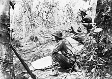 Schwarzweiss-Foto eines Mannes, der Militäruniform trägt, bewaffnet mit einer großen Waffe, die sich hinlegt und die Waffe in dichtes Buschland zielt. Zwei weitere Männer in Militäruniform hocken auf beiden Seiten des liegenden Mannes.