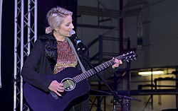 Dianna Agron, dalam mantel hangat dan gaun bermotif, memainkan gitar hitam dan bernyanyi ke mikrofon di atas panggung.