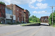several old brick buildings on the left side of a road that slopes down to a rusty railway bridge