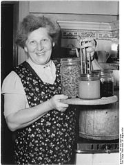 smilende eldre kvinne med krukker på gryteinnsatsen;  i bakgrunnen den tilsvarende store galvaniserte kokekaret (1954)