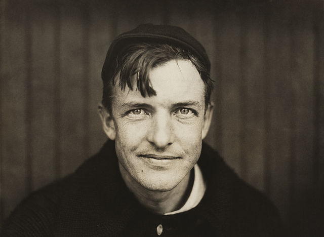 A dark-haired man wearing a black sweater and crownless baseball cap looks into the camera. His hair hangs down over his right side of his forehead, and he has a slight smirk on his face.