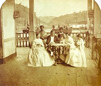 a man and three women seated around a table with two servants standing in the background