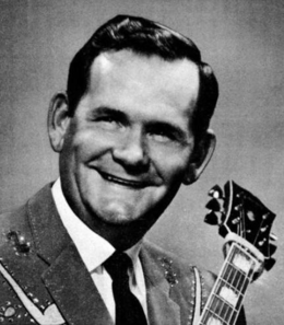 A dark-haired man in a sports jacket and tie holding a guitar.