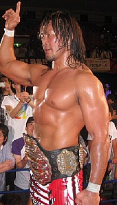 Hiroshi Tanahashi posing with the IWGP Heavyweight Championship belt in front of a crowd.