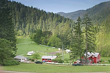 A red house and several other buildings, either red or white, are arranged on a large grassy area mostly surrounded by white fences. Two pickup trucks can be seen on a dirt road leading to one of the white buildings. Heavily forested hills rise beyond the grassy area.