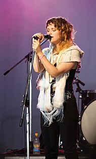 A woman with light brown hair wearing a white scarf holds and sings into a microphone