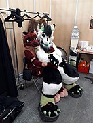 A dragon fursuiter, sitting on an otter fursuiter.