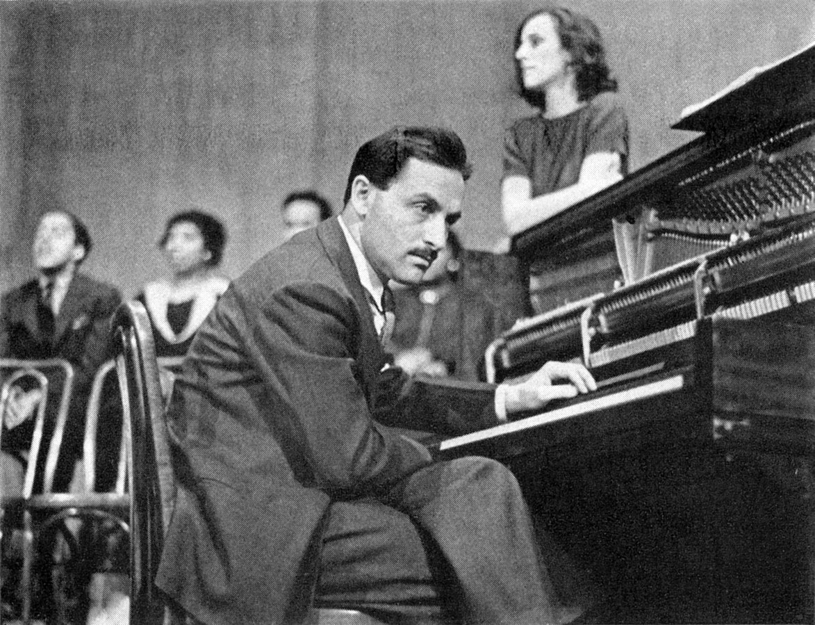 Man at open-faced upright piano, leaned a bit forward left hand on keys, turned partially toward camera. Several singers arrayed behind him in softer focus.