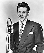 Fotografia de um jovem de terno e gravata em pé ao lado de um microfone CBS em forma de pílula em um pedestal