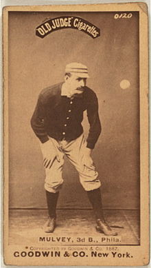 На бейсбольной карточке цвета сепии изображен мужчина в старинном черном бейсбольном свитере, белых штанах и шляпе для пилюль.