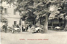 Černobílá pohlednice s restaurační zahrádkou a mnoha sedícími hosty pod košatým stromem u budovy někdejší střelnice.