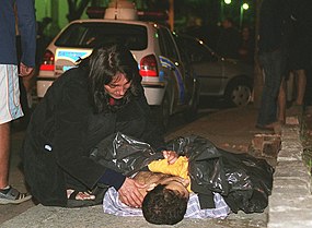 Imagem que mostra o corpo do jornalista Fernando Villela de Andrade Neto estendido na calçada com uma mulher ao lado de cócoras com a mão sobre seu ombro.