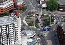 Luftaufnahme des Kreisverkehrs, einer Kreuzung mehrerer Straßen.  Fahrzeuge umfahren den Kreisverkehr, der von Gebäuden, meist mehrstöckigen, umgeben ist