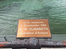 Fotografía en color de una placa de cobre que recuerda el lugar del asesinato de la emperatriz Elisabeth frente a un cuerpo de agua.