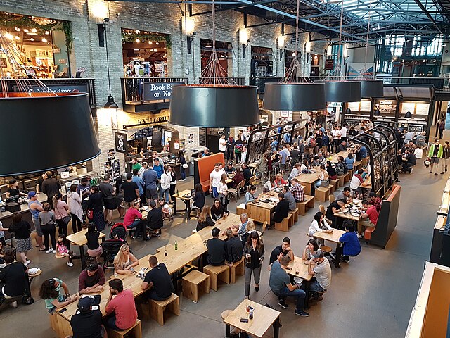 The food hall at the Forks in downtown Winnipeg, Manitoba. Photo is taken from the second level, looking down on crowded tables of people eating and d