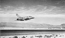 Rear quarter view of a single engine jet fighter taking off from a runway located in a desert. The bare metal finish jet has a checkerboard unit insignia on its vertical tail. The landing gear are raised. The horizon is hilly.