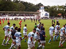 Jucătorii se antrenează pe marginea unui teren în timp ce are loc un meci de rugby.