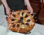 Hands holding a knife and fork in their pals support a shallow cast iron dish containing sea-food, featuring a number of medium sized crustaceans arranged in a radial pattern, heads out, interdigitated with shell fish, possibly mussels, and with lemon in five slices on top.