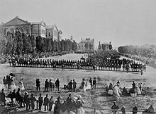 Photo en noir et blanc de militaires assemblés dans une place avec des spectateurs les observant ; en arrière plan, des bâtiments