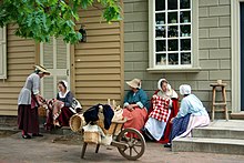 Cinco mulheres vestidas com roupas compridas de estilo colonial sentam-se nas escadas de prédios bege e bege conversando.  Na frente deles está um carrinho de mão de madeira cheio de cestas de vime.