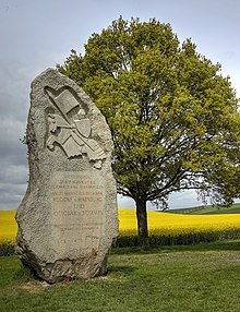 Vztyčený kámen na louce, na němž je vytesán text a vyobrazení muže se štítem a kopím s vlajkou sedícího na koni. Kůň stojí na zadních nohách. V pozadí se nachází strom a za ním řepkové pole.