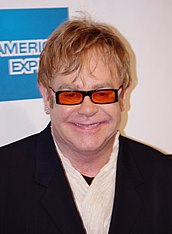 A Caucasian man with light brown hair. He is wearing black framed glasses with orange lenses and a black suit over a white shirt. Behind him is a white wall with a blue square.