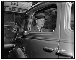 Charles Lindbergh, seen through the right-hand backseat window of an automobile. He wears a brimmed hat, a suite, a white button-up shirt, and a dark-colored necktie. His expression is serious.