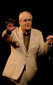 Photographie couleur d'un homme vieillissant, au front dégarni, qui debout sur une scène salue un public.