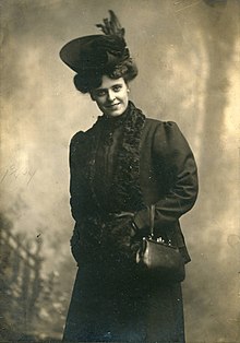 A white woman, standing and smiling, wearing a dark hat, coat, gloves, and holding a dark handbag over one arm.